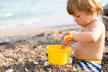 Cute child playing with yellow bucket and toy car on pebble beach. Happy toddler on sea coast. Summer family holiday vacation. Two years old little boy having fun. Summertime. Travel concept