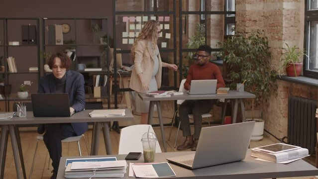 Timelapse of long-haired Caucasian expecting woman using laptop, drinking green juice, talking to diverse colleagues in modern coworking space in afternoon