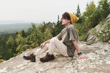 Dreamy young Caucasian hiker in yellow hat and boots sitting on big stone and contemplaing forest landscape