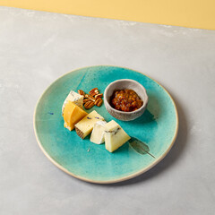 Cheese platter on blue ceramic plate. Cheeseboard on handmade dinner plate in modern style. Different cheese on ceramic dishware in minimal style.