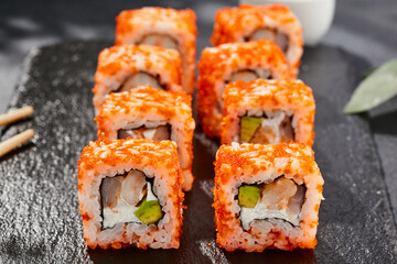 Maki sushi on dark stone table. California maki with tobiko. Sushi roll with cheese,shrimp, avocado inside, maguro outside. Style concept japanese menu with black background, leaves and hard shadow.