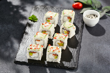 Fresh maki sushi on dark stone table. Colorful roll with vegetables. Sushi roll with salmon, avocado inside, cucumber outside. Style concept japanese menu with black background, leaves and shadow.