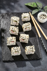 Maki sushi on dark slate. California maki with sesame. Sushi roll with crab, cucumber inside, sesame outside. Style concept japanese menu with black background, leaves and hard shadow.