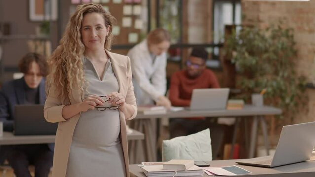 Portrait of curly blond-haired Caucasian mother-to-be in formalwear standing in modern office at daytime, looking and smiling on camera