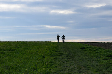 Fototapeta premium Silhouettes of a running man and a cyclist side by side against a cloudy blue sky on the horizon line