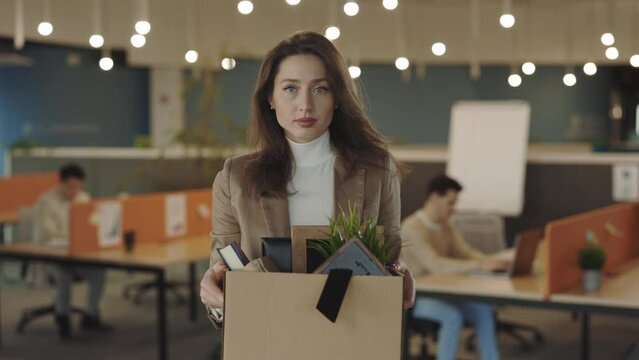 Portrait of jobless caucasian female emloyee fired from work. Upset young woman with box full of things in hands looking at camera. Indoors. Firing from job. Crisis and business