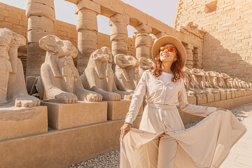 A happy tourist girl interested in Egyptology and archaeology and gets a travel experience at the Karnak Temple in Luxor among alley of sphinxes in Thebes - obrazy, fototapety, plakaty