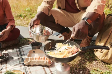 Foto op Canvas Close-up of unrecognizable black man in smartwatch cooking eggs on burner and putting it in metallic bowl outdoors © Mediaphotos