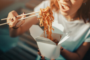 Happy Asian girl holds chopsticks and eats wok tasty noodles from a takeaway paper box. Street and...