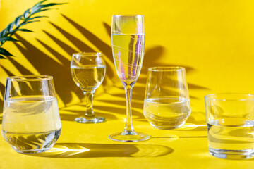 A various glasses of mineral water on yellow background with green leaves and hard shadows.