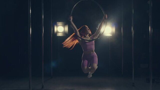 In the dark, in the backlight, a red-haired girl in a pink suit holds her hands on a gymnastic hoop and rotates smoothly on it, the girl gracefully bends with each turn of the hoop and changes her