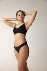 Obraz premium Portrait of young woman with natural body posing in black underwear isolated over grey studio background. Femininity