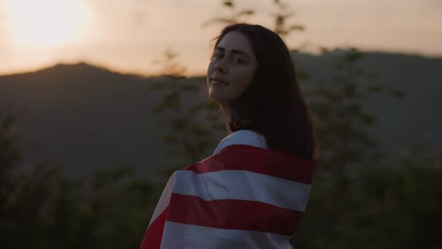 Independence Day. A young woman with an American flag draped over her shoulder and turn around, against the background of the sunset sky. Slow motion. The concept of patriotism.