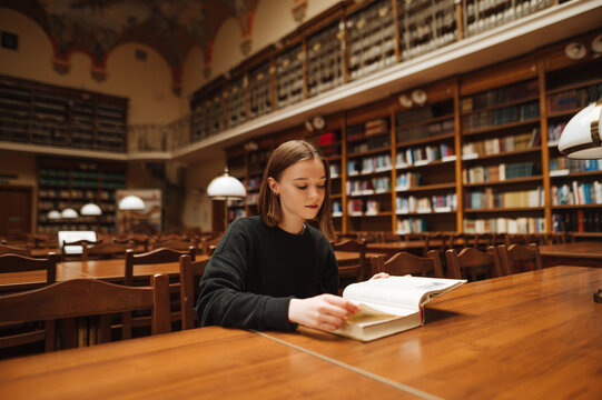 Girl student in dark clothes studies at home with a book on the table in the public library.