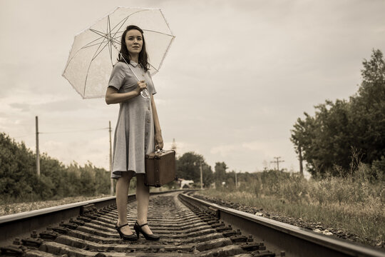 A young fashionista woman in vintage style with a suitcase and a summer umbrella stands on the railway and looks ahead purposefully.