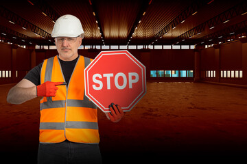 Builder warns of danger. Inspector in orange vest demonstrates stop sign. Concept of danger of using industrial building. Security inspector points to word Stop. Builder inside dark hangar. - obrazy, fototapety, plakaty
