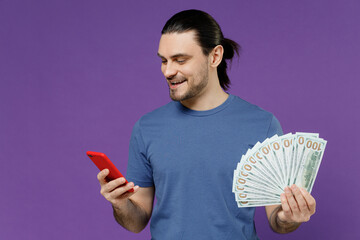Young smiling man 20s wear basic blue t-shirt hold mobile cell phone holding fan of cash money in...