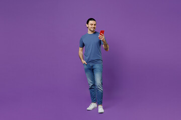 Full body young smiling man 20s in basic blue t-shirt hold in hand use mobile cell phone chatting online browsing isolated on plain purple color background studio portrait. People lifestyle concept.