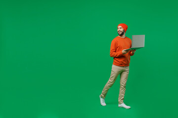 Full body young smiling happy man 20s in orange sweatshirt hat hold use work on laptop pc computer...