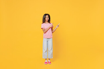 Full body fun little kid girl of African American ethnicity 12-13 years old in pink t-shirt point index finger adide on workspace area isolated on plain yellow background. Childhood lifestyle concept.