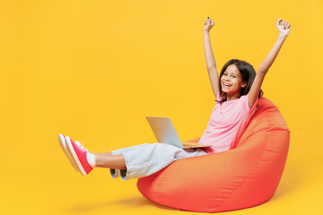 Full body fun little kid girl of African American ethnicity 12-13 years old in pink t-shirt sit in bag chair hold use work on laptop pc computer stretch hands legs isolated on plain yellow background