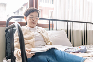 Portrait of senior Asian woman wearing sweater sitting on the sofa and enjoying reading a magazine at home.