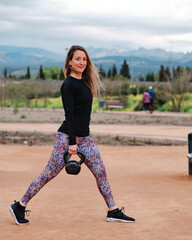 Young blonde woman performing calisthenics exercises in an urban park. She is doing kettlebell weight lifting exercises.