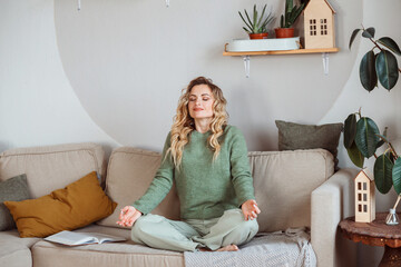 Good looking woman in cosy green sweater sitting on the sofa and meditating at home.
