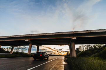 Beautiful nature landscape with a traveling car on a wet asphalt road after rain with a concrete bridge against the backdrop of a sunset and a blue sky with clouds.