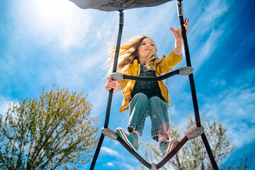 A young girl in a yellow raincoat climbs up a rope ladder with a bright blue sky in the background.