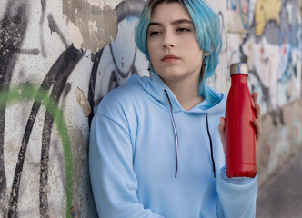 Blue haired Teenage girl in blue hoodie staying near graffiti wall with red water bottle