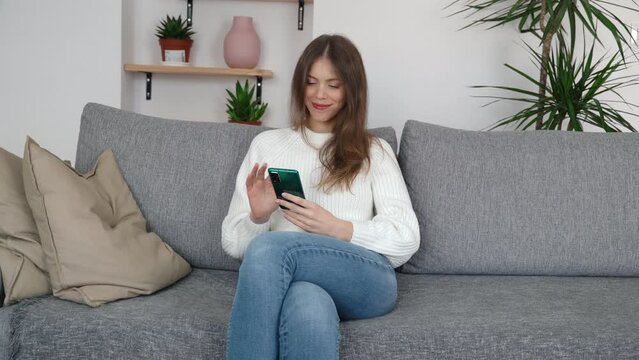 A young girl sits in a relaxed pose on a sofa in a modern room with a phone in her hands and looks at the screen and smiles.