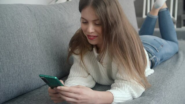 A beautiful young girl lies on the couch with a modern smartphone in her hands and looks through the content on the screen and smiles.