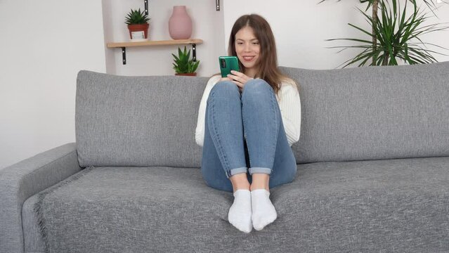 A young girl is sitting in a relaxed position on the sofa with her legs up and a phone in her hands, she is typing something on the screen and smiling.