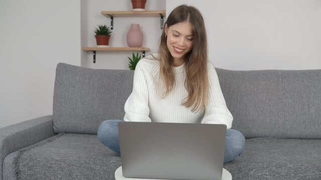 A young girl sits cross-legged on a sofa in a modern room and works on a computer. The concept of remote work or study.