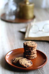 Stack of chocolate chip cookies, open book and reading glasses, lit candle and vase with flowers on the table. Hygge at home. Selective focus.