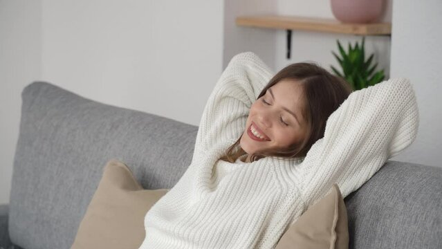 A young girl sits on a couch with her hands behind her head and closed eyes. The concept of rest in the middle of the day. Side view.