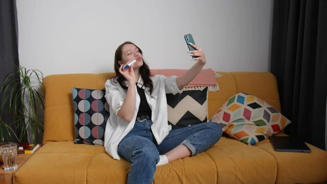 A young woman is sitting on the couch and taking a selfie on her phone with a pregnancy test. A young woman is pregnant. Early pregnancy