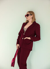 teen fashion. young blonde girl in vinous classic suit and sunglasses and pink bag in hand is standing fashion and looking apart on the white wall background in studio. fashion concept, free space