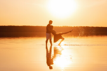 Silhouette of a couple in love, dances with splashes in the water of the lake in the orange sunset