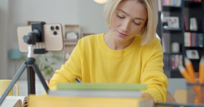 Charming young woman sitting at desk with modern cell phone that fixed ib tripod and having online lesson. Concept of people, technology and distance learning.