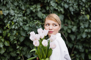 Fototapeta premium young blonde woman with white tulips smiling near green ivy outdoors.