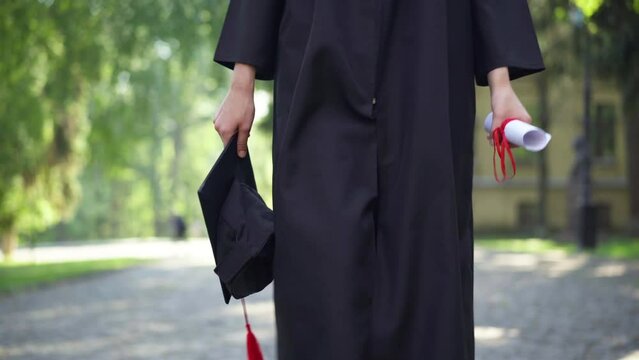 Dolly shot of unrecognizable slim young Caucasian woman in graduation toga walking at university campus. Front view confident intelligent graduate student with diploma strolling outdoors