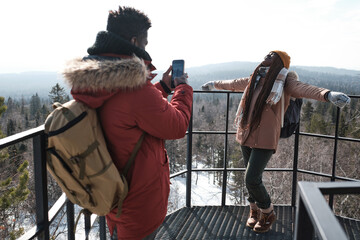 Young African American guy standing at viewing platform on mountain top taking pictures of his girlfriend on smartphone camera