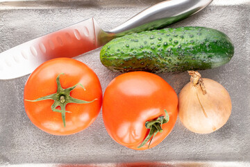 Two ripe tomatoes, a cucumber and an onion with a knife on a metal tray, macro, top view.