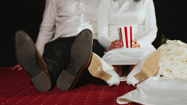 Married couple having fun alone in movies. Elegantly dressed man and woman in wedding clothes sit on floor of cinema and wave feet in shoes. Bride holds box with popcorn. Wedding bouquet near them