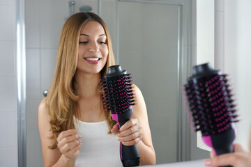 Smiling girl holds round brush hair dryer in her bathroom at home. Young woman looking satisfied her salon one-step brush hair dryer and volumizer.