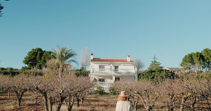 Woman walks between bare trees towards big two-storey house, look behind