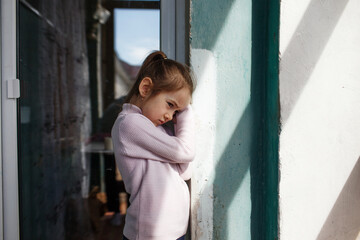 a sad little girl is standing near the window.