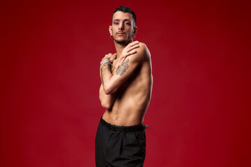 Fototapeta na wymiar Young model with tattoos, shirtless and makeup. Posing with a red background, showing his feelings.